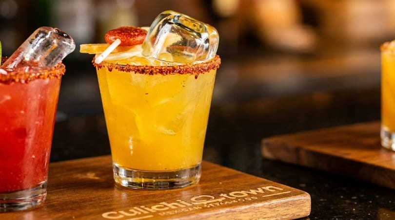Two vibrant cocktails, one red and one yellow, garnished with spices and fruit on a wooden board.