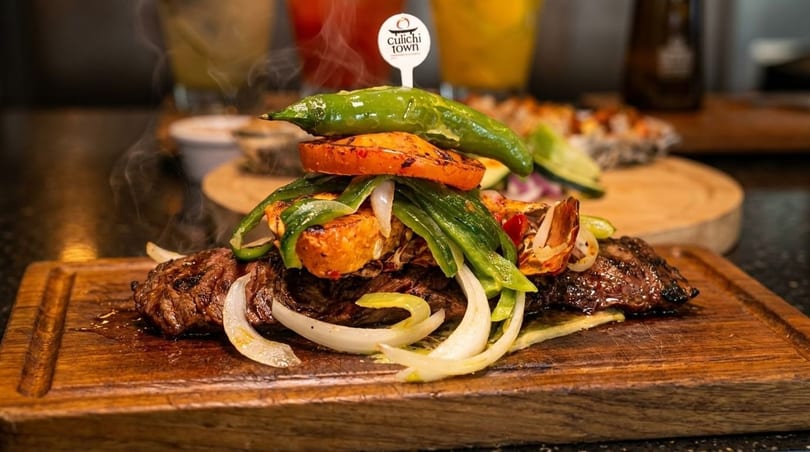 Steaming grilled steak with onions, peppers, and a Culiichi Town pick on a wooden board.