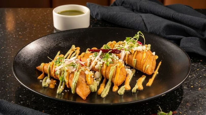 Crispy fried taquitos drizzled with green and orange sauces, topped with cheese, radishes, and herbs.