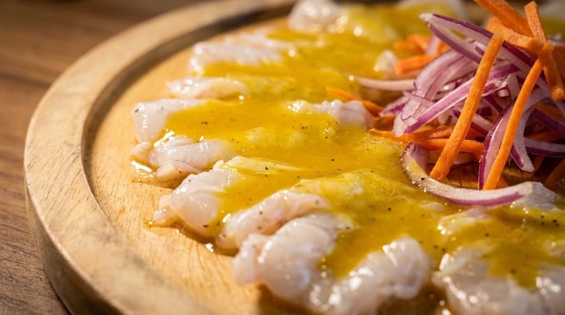 White fish ceviche with yellow sauce, julienned carrots, and red onions on a wooden plate.