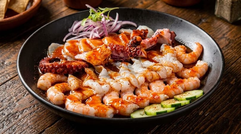 A black bowl filled with shrimp, octopus, and fish ceviche, garnished with red onion and cucumber.