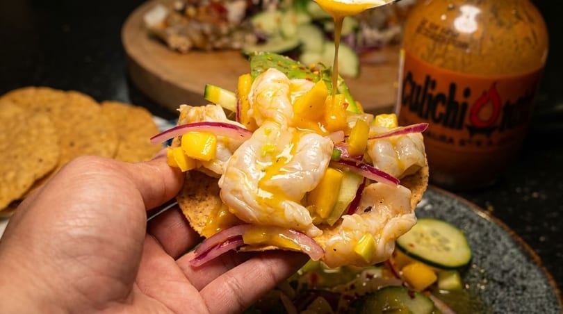 A hand holds a shrimp tostada topped with mango, red onion, and sauce drizzling from above.