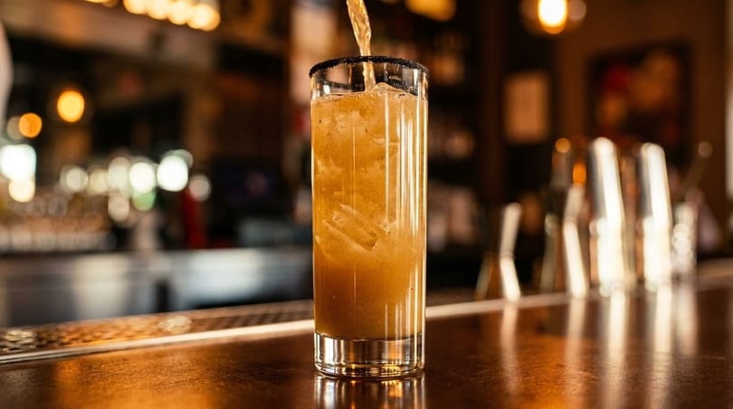 A tall glass with a dark rim, filled with a brown drink and ice, on a bar.