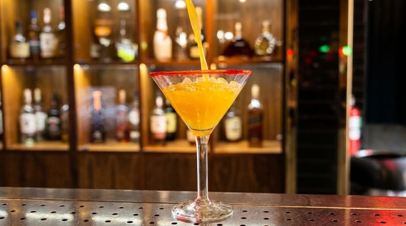 A bright orange cocktail with a red sugar rim is being poured into a martini glass at a bar.