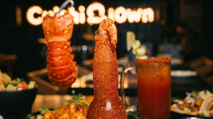 A spicy shrimp cocktail bottle and a lobster tail skewer on a dark restaurant table.